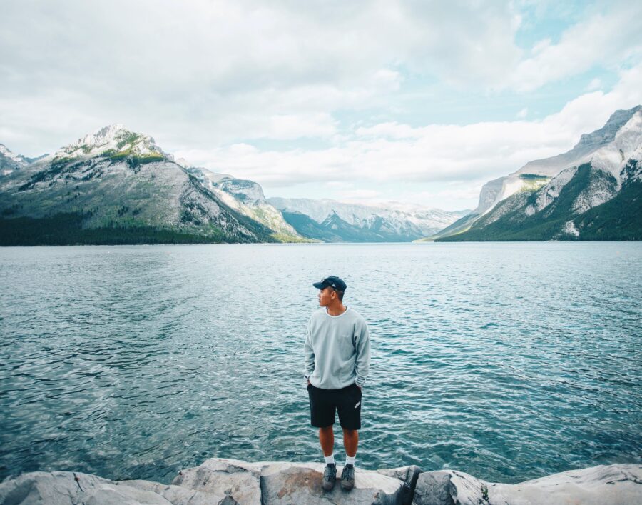 A Man in Gray Sweater Standing Near the Body of Water while Looking at the Mountain
