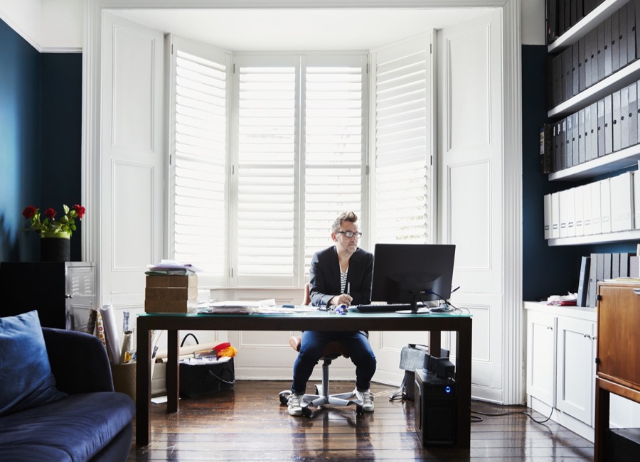 furniture designer at shop and home,united kingdom,a man in suit and trainers with glasses, sitting at a desk in a bay window. a light airy office with files on shelves.